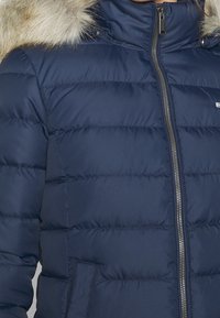 Mörkblå vadderad vinterjacka med frontdragkedja och huva med pälsfoder, bärs av en person med synlig nacke och haka.