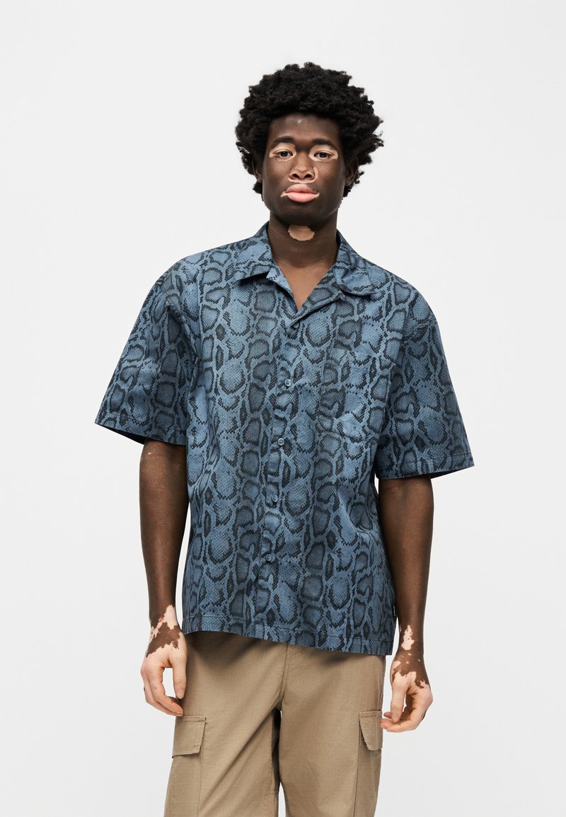 Camisa de manga corta con botones en azul con un estampado de serpiente negra. Fabricada con tela suave, presenta un corte relajado y un bolsillo.