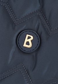 Navy materiał pikowany z wyraźnym okrągłym logo w czerni z złotym "B" w środku; ukośne szwy dodają teksturalnego detalu.