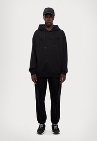 Sudadera negra con logo en relieve, combinada con pantalones negros que presentan un patrón de olas. Zapatillas negras completan el conjunto.