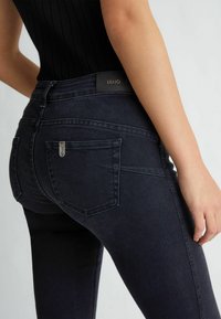 Jeans de denim oscuro de tiro alto con una forma ajustada, que presentan bolsillos traseros y una etiqueta de marca de cuero en la cinturilla. Textura suave.