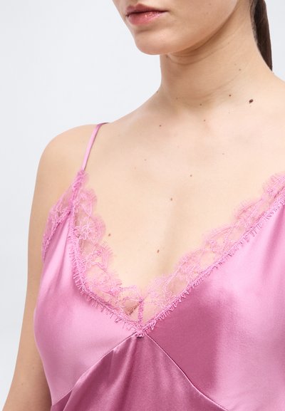 Γυναίκα που φοράει ροζ σατέν καμιζόλ με φλοράλ δαντέλα κατά μήκος του V-λαιμού και λεπτούς ιμάντες στους ώμους.