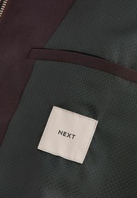Gros plan sur un tissu sombre avec un motif hexagonal, une bordure bordeaux et une étiquette blanche rectangulaire portant la mention « NEXT ».