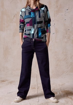 Femme portant un haut à manches longues à motifs géométriques multicolores, jean large foncé, et baskets blanches, debout les mains dans les poches.