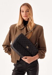 Borsa a tracolla in pelle trapuntata nera con catena e un accento in metallo con il logo. La borsa presenta una forma rettangolare e una texture liscia.