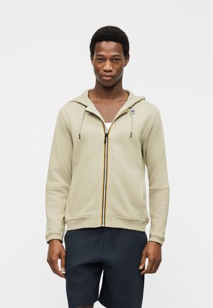 Moški, ki nosi bež kapucar z zadrgo čez belo majico in temne kratke hlače, stoji pred enobarvnim belim ozadjem.