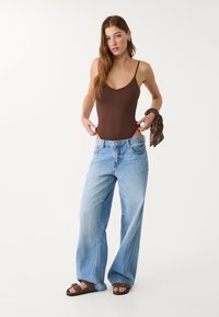 Body in cotone senza maniche marrone abbinato a jeans larghi di un blu sbiadito. La modella indossa sandali marroni e tiene un accessorio a sciarpa marrone.