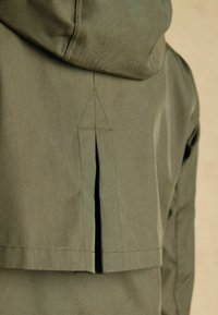 Zielona oliwkowa kurtka z kapturem, z trójkątnym detalem szwu i wentylacją z tyłu, wykonana z gładkiego materiału o subtelnym połysku.