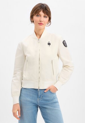 Donna che indossa una giacca bomber color crema con tasche con cerniera e toppa sulla manica sinistra, abbinata a jeans azzurri, su uno sfondo bianco.