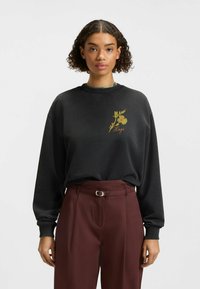 Svart sweatshirt i mjukt tyg, med en blommig design i grönt och gult på bröstet. Bärs med vinröda högmidjade byxor.