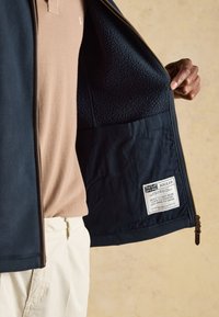 Person hält eine offene marineblaue Fleecejacke, die eine Innentasche und ein Etikett mit dem Markennamen "Joules" und Text über Handwerkskunst zeigt.