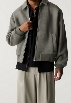 Personne portant une veste grise zippée sur une chemise noire et un pantalon beige, montrant des bagues et des colliers en argent aux mains et au cou.