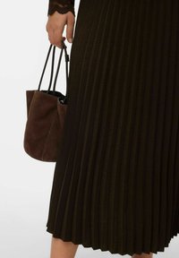 Sac seau en daim brun tenu par une main, contrastant avec une jupe plissée foncée. Le tissu a une apparence texturée et côtelée.