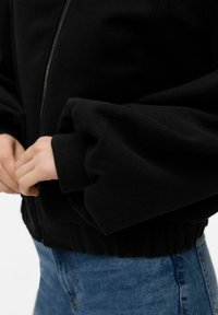 Veste noire zippée avec une texture lisse, dotée de manches bouffantes et d'un ourlet froncé. Associée à un jean en denim bleu.