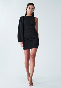 Melna pieguļoša kleita ar vienu garu piedurkni, ar ručkotu tekstūru, virs ceļa garumā, kopā ar zelta krāsas sandales ar siksnām un lielām apaļām auskariem.