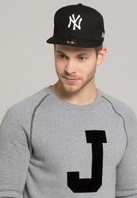 Černá baseballová čepice s bílým logem "NY", nošená s šedým svetrm, na kterém je vpředu velké černé "J". Jednoduché pozadí.
