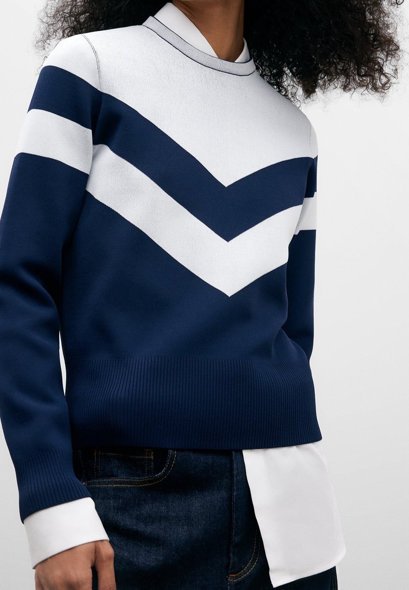 Pull bleu marine avec des rayures en chevrons blanches, ourlet et col côtelés. Porté par-dessus une chemise blanche, offrant un look à couches superposées.