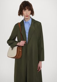 Bruin en crème schoudertas met een gladde textuur, rechthoekige vorm en gouden accent. Draagt bij een olijfgroene jas en blauwe blouse.