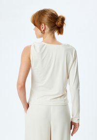 Top à manches longues en tissu écru, avec un design drapé et des manches asymétriques. Texture lisse avec des détails de couture subtils.