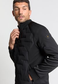 Zwarte jas met een gevoerde voorkant en een gladde achterkant, hoge kraag, ritssluiting en een hexagonaal logoaccent op de mouw.