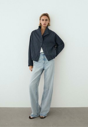 Vrouw draagt een donker gestreepte jas over een wit topje, lichtblauwe wijd gesneden jeans en zwarte sandaal met open teen, staand tegen een witte muur.