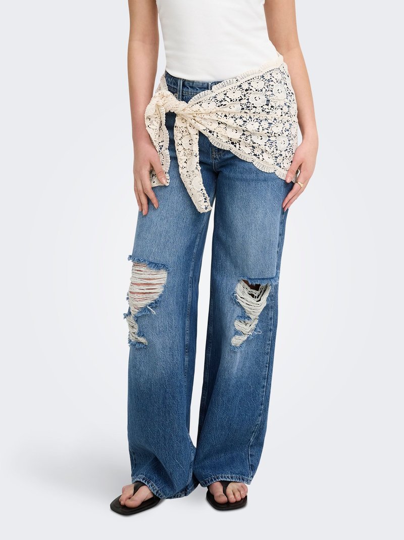 Mujer con vaqueros azules anchos y rotos, camiseta blanca, pañuelo de encaje beige atado a la cintura y chanclas negras, de pie frente a un fondo liso.