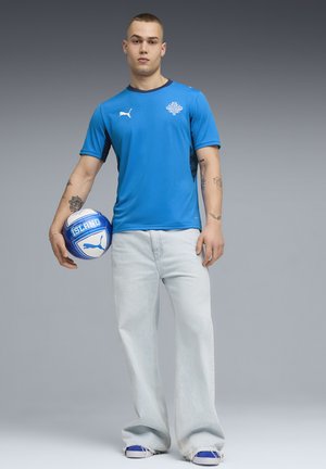 Μπλε αθλητική κοντομάνικη μπλούζα με μαύρες λεπτομέρειες και λογότυπο, συνδυασμένη με ανοιχτόχρωμο τζιν παντελόνι και μπλε αθλητικά παπούτσια, κρατώντας μια μπλε ποδοσφαιρική μπάλα.