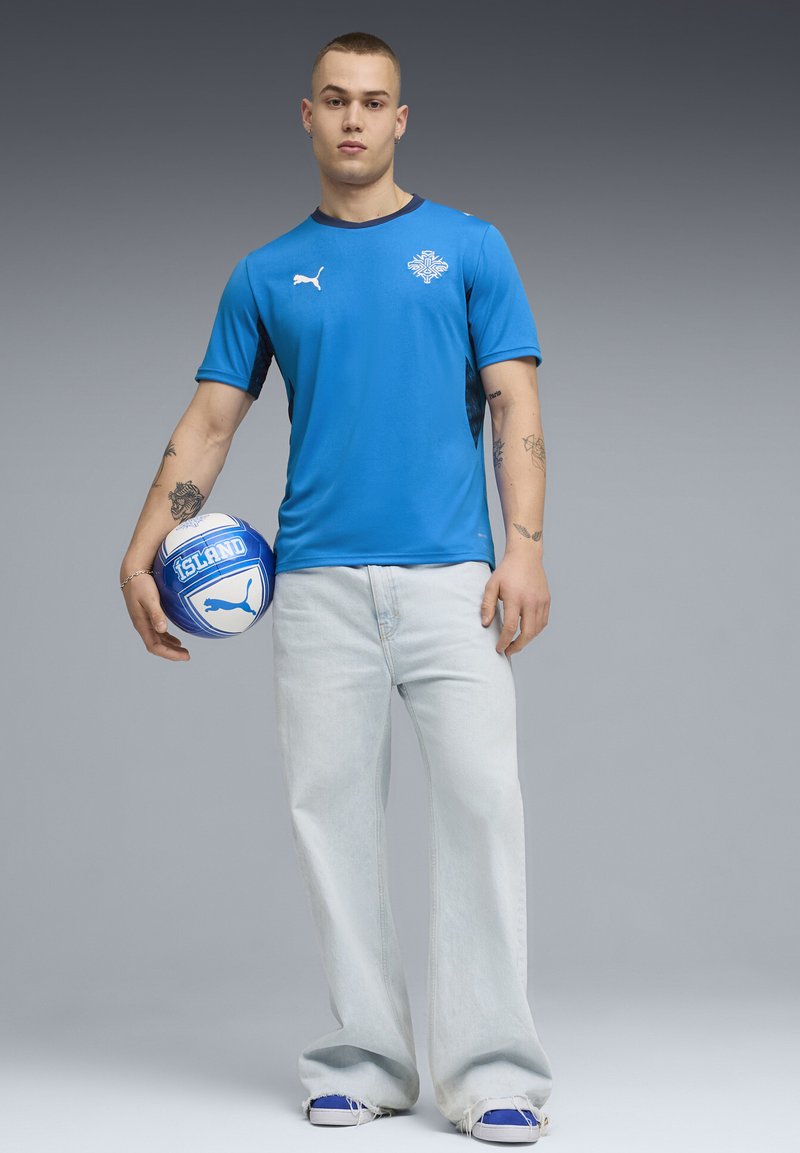 Μπλε αθλητική κοντομάνικη μπλούζα με μαύρες λεπτομέρειες και λογότυπο, συνδυασμένη με ανοιχτόχρωμο τζιν παντελόνι και μπλε αθλητικά παπούτσια, κρατώντας μια μπλε ποδοσφαιρική μπάλα.