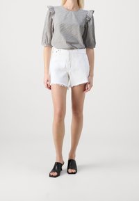 Una donna indossa una blusa a maniche puff a scacchi, pantaloni corti bianchi con orlo sfrangiato e mule aperte nere con tacco, in piedi davanti a uno sfondo neutro.