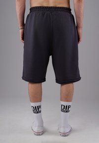 Schwarze Sportshorts mit einem elastischen Bund, bis zur Mitte des Oberschenkels. Kombiniert mit weißen Socken mit schwarzer Schrift und weißen Sneakern.