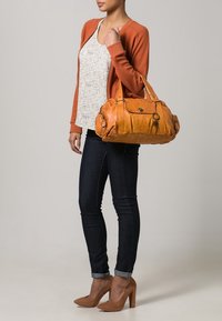 Sac à main en cuir tan de forme arrondie, avec deux anses et des détails métalliques discrets. Porte avec un jean foncé et un cardigan orange.