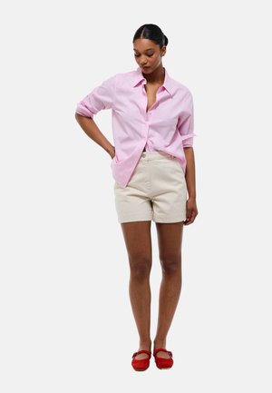 Femme portant une chemise rose clair à boutons, un short beige et des chaussures plates rouges, debout, une main sur la hanche, regardant vers le bas.