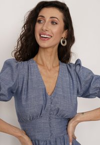 Jasnoniebieska sukienka lniana z dekoltem w serek, bufiastymi rękawami i guzikami z przodu. Gładka tekstura z dopasowanym pasem i rozkloszowanym dołem. Złote kolczyki w formie kółek.