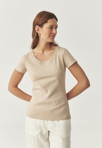 T-shirt beige à manches courtes avec un col rond, fabriqué à partir d'un tissu doux. Présente une silhouette ajustée et des détails de couture subtils.