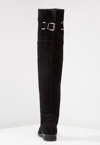 Botas de ante negras por encima de la rodilla con una textura suave, que presentan un acento de hebilla plateada en el costado y un tacón plano.