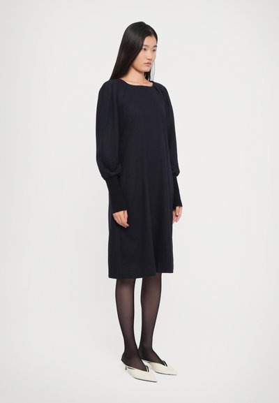 Woman standing wearing a long-sleeve black dress with puffed shoulders, black tights, and white pointed-toe heels against a plain background.