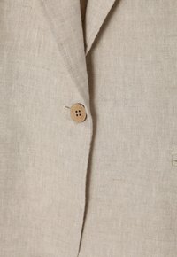 Beige vászon blézer texturált anyagból, egyetlen kerek faszegély gombbal és kiemelt hajtókával. Minimalista dizájn, tiszta vonalakkal.