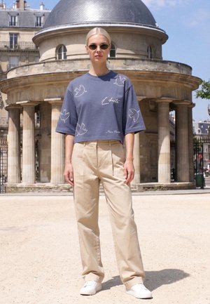 Blonde woman wearing sunglasses, a blue shirt with white bird prints, beige pants, and white sneakers stands in front of a historic stone building with columns.