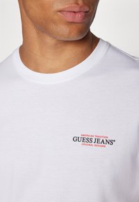 Bílý bavlněný tričko s kulatým výstřihem a černou a červenou logem "GUESS JEANS" na hrudi, s dalším textem nad a pod logem.