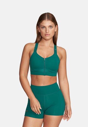 Kobieta nosząca sportowy biustonosz na zamek w kolorze morskim i dopasowane wysokie szorty, stojąca na jednolitym jasnym tle.