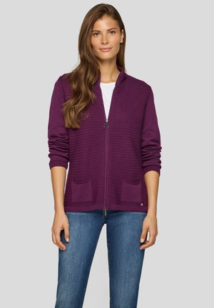 Lila gerippter Cardigan mit einem Reißverschluss, zwei Vordertaschen und einem Stehkragen, kombiniert mit einem weißen Oberteil und blauen Jeans.