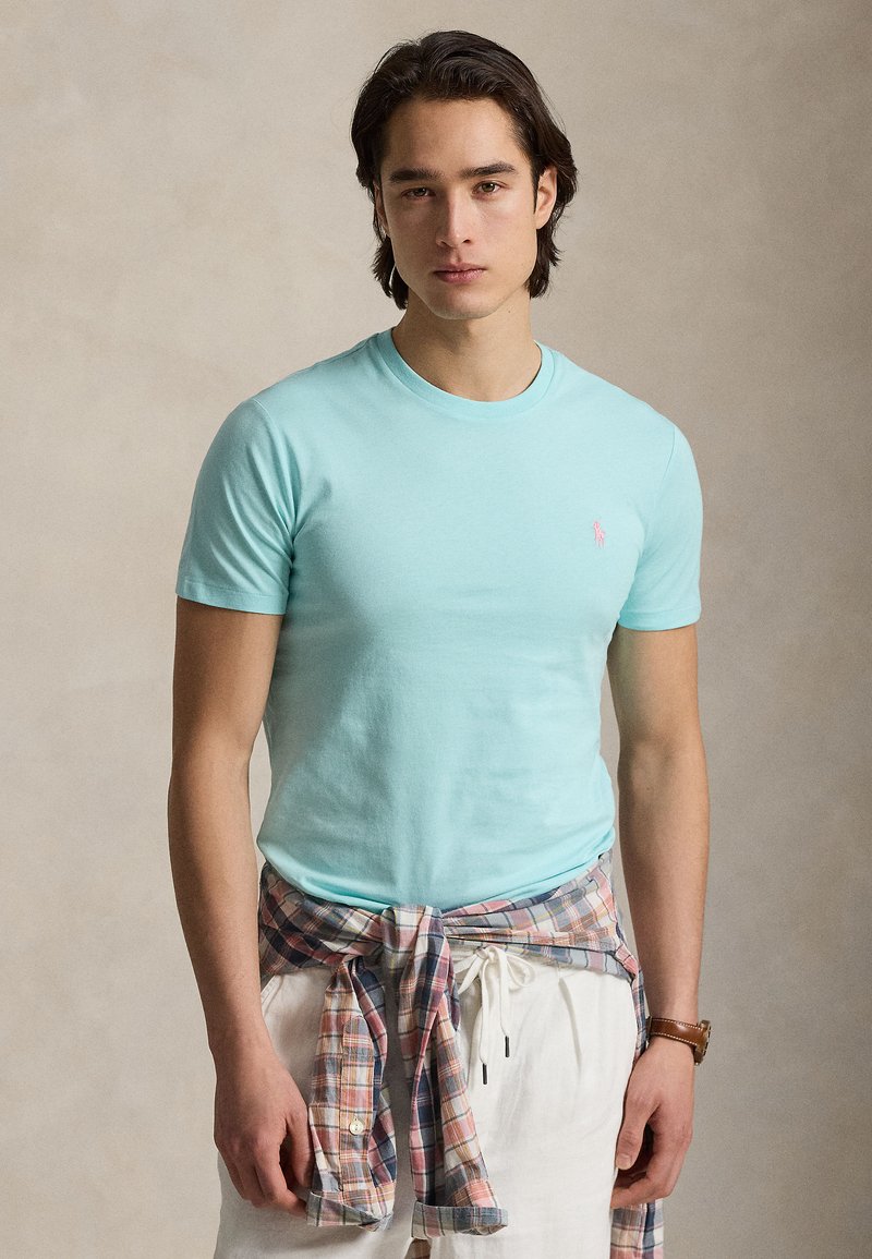 Νέος άνδρας με ανοιχτό γαλάζιο t-shirt με ροζ λογότυπο, καρό πουκάμισο δεμένο στη μέση του, άσπρο παντελόνι και καφέ ρολόι χειρός.