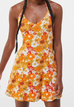 Kvinde iført en ærmeløs, V-udskåret blomsterkjole med orange, gule, hvide og røde blomster, stående mod en ensfarvet baggrund.