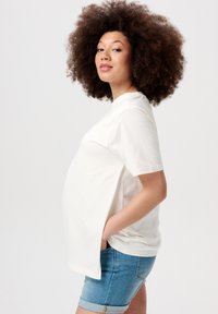 Hvid kortærmet skjorte lavet af blødt bomuld, med sideskår, parret med lyseblå denimshorts og set fra siden.