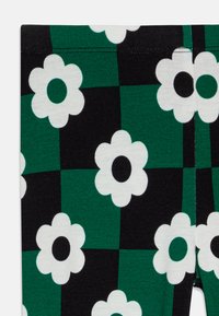 Leggings con un patrón de tablero de ajedrez en negro y verde, decorados con grandes flores blancas y centros negros. Tejido suave y elástico.