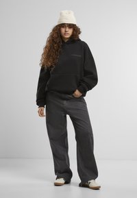 Schwarzer übergroßer Hoodie mit einer Fronttasche und dezentem Branding, kombiniert mit dunkelgrauen weiteren Jeans und cremefarbenen Sneakers.