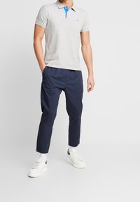 Homme portant un polo gris clair avec un col bleu, un pantalon court bleu marine, des chaussettes blanches et des baskets blanches, debout les mains dans les poches.