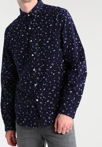 Námořnicky modrá košile s dlouhým rukávem, zdobená rozptýleným bílým vzorem různých malých tvarů. Košile má límec na knoflíky a zapínání vpředu na knoflíky.