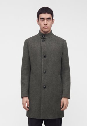 Man with short dark hair wearing a buttoned, high-collar gray textured coat standing against a plain white background.