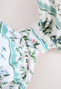 Prim-plan al unui material cu model floral în verde, roz și albastru, cu secțiuni elastice strânse, pe un fundal alb.